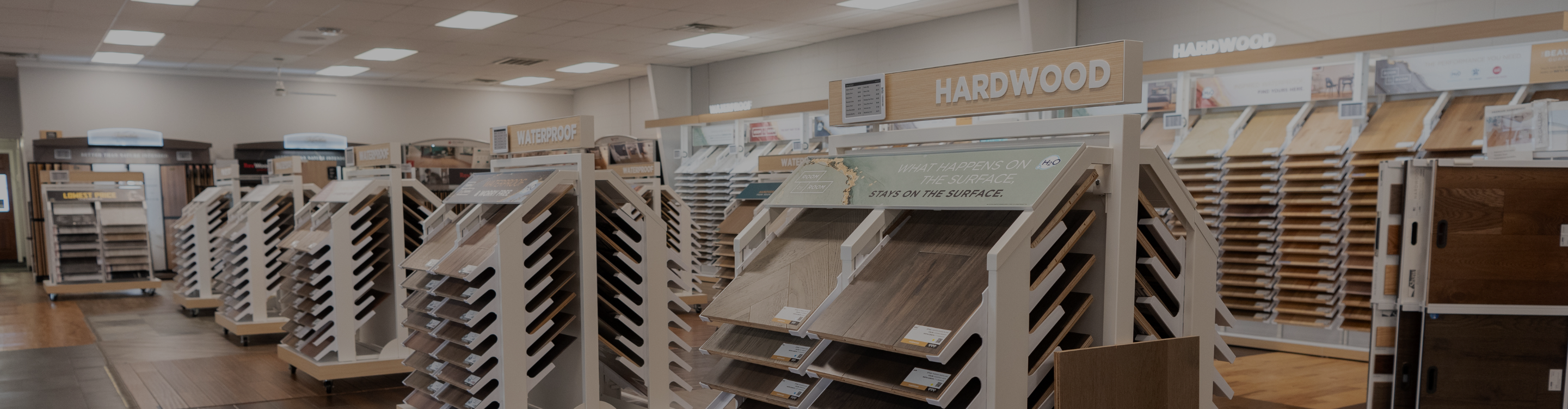 Carpet One of Spartanburg showroom aisle displaying various flooring samples, including hardwood and laminate, arranged on tiered racks under "HARDWOOD" signage.